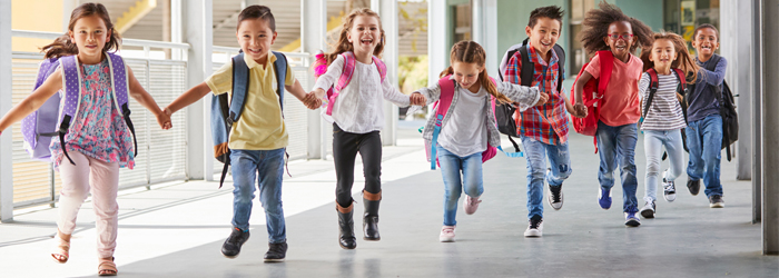 Crianças na volta às aulas, de mãos dadas, sorrindo e com mochila nas costas.